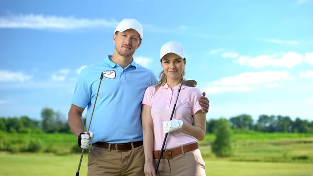 Attractive couple of golfers with clubs posing on camera at course, sport school