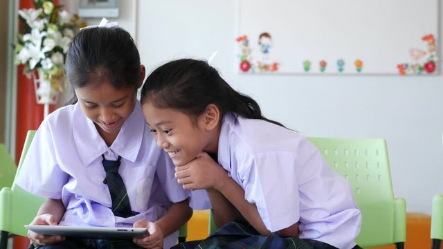 Two Thai cute elementary schoo lgirls, long hair in a white shirt dress, skirt and green plaid tie are playing games on the tablet easily in school.