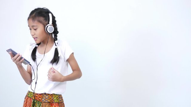 The girl looks cute and wears a white shirt and a braid tied with a white ribbon using the headphones, listening to music from social media on smartphone, singing softly and rocking along the song.