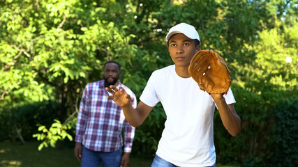 Attentive black teenager playing baseball in park, dad supporting son, sport