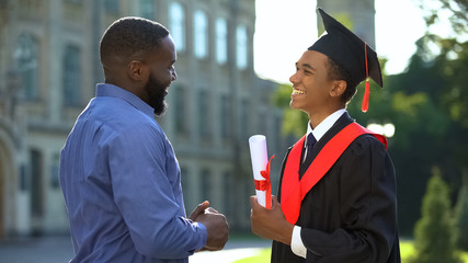 Happy proud man greeting teenage son in graduation gown, educational success