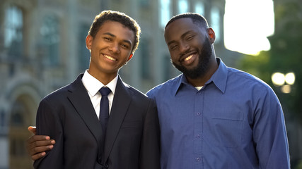 Joyful dad hugging smart graduating son suit smiling camera, educational success