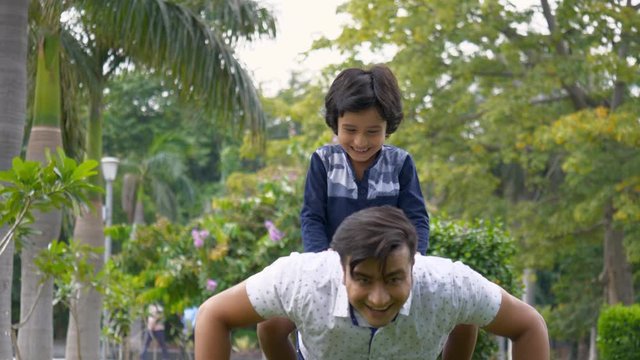 Smiling father and young son spending the weekend together in a park - leisure concept. Attractive Indian man giving push-ups ride to his cute kid sitting on his back - Indian parenting lifestyle