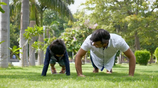 Indian father teaching his young son how to do pushups in a park for a healthy lifestyle. Attractive dad and child training together while doing gymnastic exercises on a Sunday morning - Push-ups e...
