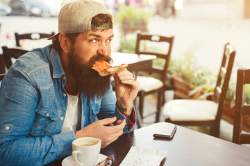 Handsome bearded man eating a slice of pizza outdoor terrace. Hipster guy has a break time for...