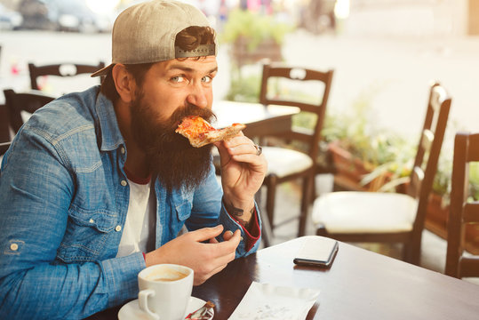 Handsome bearded man eating a slice of pizza outdoor terrace. Hipster guy has a break time for coffee and lunch. Street style, lifestyle