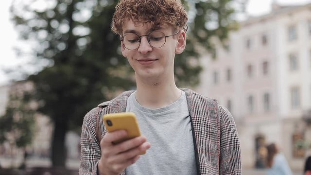 Young Nerdy Student in Glasses Using his Modern Mobile Phone Device, Scrolling on the Screen Smiling Walking Outdoor. Communication, Student, Tourist Concept.