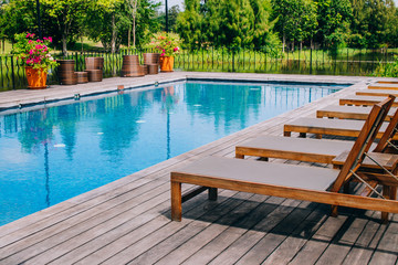 Wooden daybed place on wooden floor beside swimming pool in the resort with green natural in background.