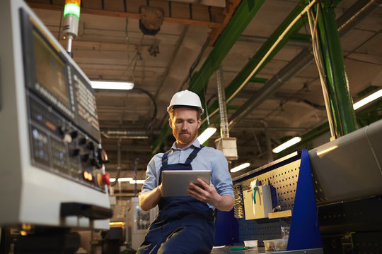 Young operator in protective work wear using digital tablet while standing near the machine in the factory