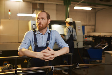 Serious bearded manual worker in overalls standing near the lathe of production metal pipes with his colleague in the background in the factory