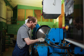 Young mechanic in work wear standing near the lathe and repairing it in the plant