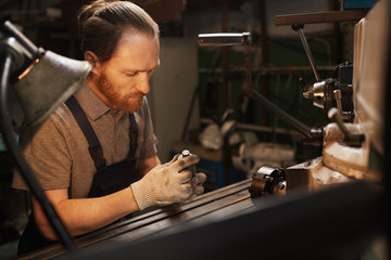 Serious bearded mechanic working with metallic objects near the lathe in the factory