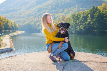 Young attractive blonde woman and her German Rottweiler dog sitting on background of beautiful autumn landscape. Friendship, pet and woman