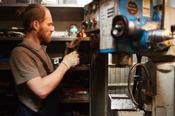 Young bearded mechanic examining metal details while working in warehouse