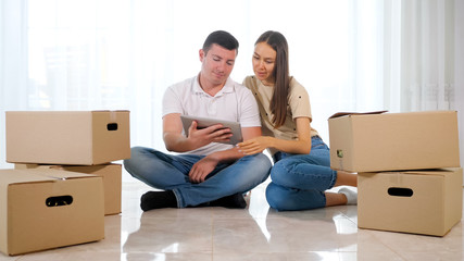 couple wife husband plans furniture location in new house sitting on floor among big cardboard boxes and uses tablet