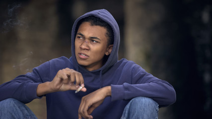Difficult teenage boy smoking cigarette sitting street, lack of parental control