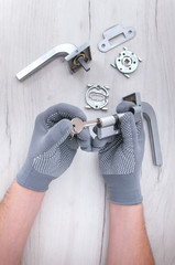 The guy holds in his hands in gray gloves the core of the lock and door keys on a light background. Repair in the house. Installing a new lock on the door. Spare parts for the door on a gray wooden ta