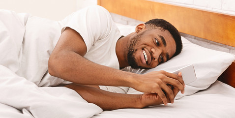 African american guy chatting with friends on smartphone in bed