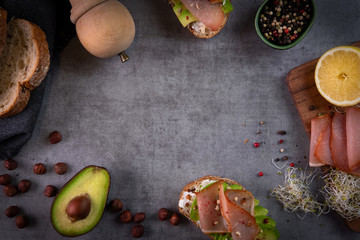 Making avocado sandwiches flat-lay on dark grey stone slate background. Healthy breakfast, healthy food concept. Avocado toast with goat cheese, nuts, sprouts, ham and spices. Top view, copy space