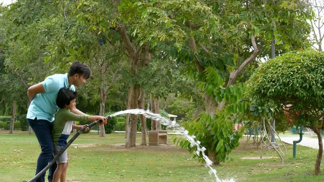A young man and his kid sprinkling water over big trees in a garden - leisure time. Attractive Indian father and his little son having fun in watering trees using a black water pipe - ecology conce...