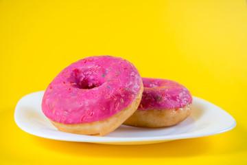 Two sweet pink donuts on a white plate isolated on a yellow background. Bright tasty bun. Minimal summer concept. Flat lay. The concept of unhealthy eating and weight gain. Harmful fast food
