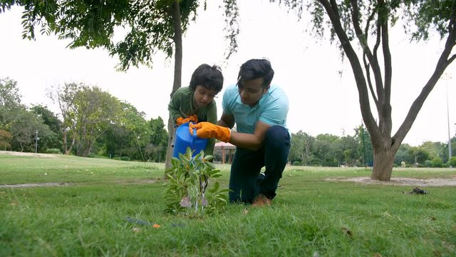 Indian father and son watering a plant while gardening in a park - save environment concept. A young man and his kid having fun in sprinkling water on a sapling with a watering can - leisure concept