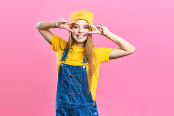 Portret a pretty girl in denim overalls and a yellow hat on a pink background. Fashionista lady student smiling . Bright trendy studio fashion image of sexy model, wearing neon bright color block