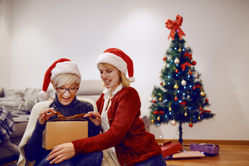 Amazed senior woman opening christmas present while sitting on floor in living room. Her daughter hugging her and sitting next to her. Both have santa hats on heads. In background is christmas tree.