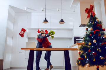 Pregnant daughter hugging her mother while standing in kitchen at home. Both having santa hats on heads. Senior woman holding lettuce. On kitchen counter is laptop. In foreground is christmas tree.