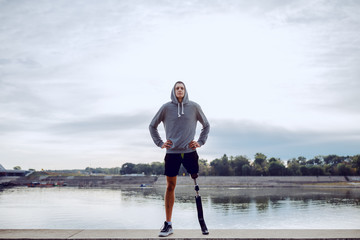 Fit healthy Caucasian handicapped man in sportswear and with artificial leg standing on quay with hands on hips.