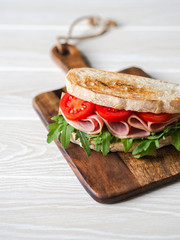 Ham, tomato and arugula sandwich on toasted bread slices on a wood cutting board on wood white background. Copy space