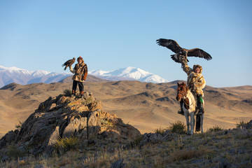 Two old traditional kazakh eagle hunters posing with their golden eagle in the mountains. Ulgii, Western Mongolia. - obrazy, fototapety, plakaty