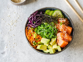 Hawaiian salmon poke bowl with rice, avocado, cucumber, carrot, red cabbage, chukka and sesame seeds on a gray background. top view