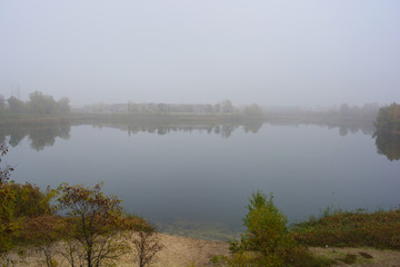Fototapeta premium Beautiful landscape, autumnal nature with white dense fog over Career Lake, Dnipro city, Ukraine. Tall trees and large shrubs with yellow leaves around a clean and beautiful lake.