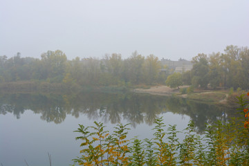 Beautiful landscape, autumnal nature with white dense fog over Career Lake, Dnipro city, Ukraine. Tall trees and large shrubs with yellow leaves around a clean and beautiful lake.