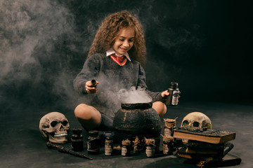 Little witch girl with long hair, dressed in dark clothes, is sitting near a steaming pot against a black studio background and preparing a potion.