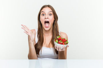 Young caucasian woman holding a strawberries bowl celebrating a victory or success