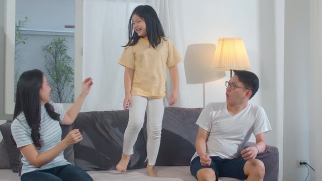 Happy young Asian family listen to music and dancing on sofa at home. Chinese mother father and child daughter are enjoying happy spending time together in modern living room in evening. Slow motion.