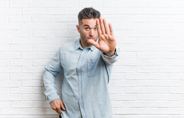 Young handsome man against a bricks wall standing with outstretched hand showing stop sign, preventing you.