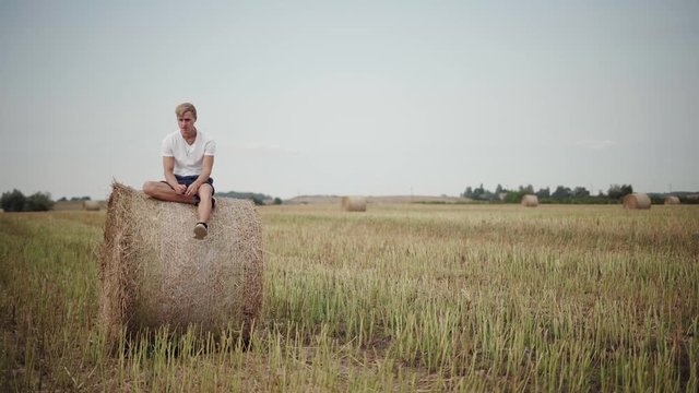 Young male relaxing by sitting on top of a hay bale in the middle of a field. Static wide shot