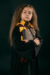 Portrait of a little witch girl with long brown hair dressed in dark coat, holding magic wand in her hand, posing on black studio background.