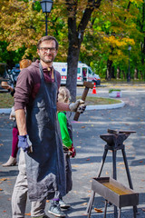 A blacksmith forges a metal blank on the anvil at a fair in the presence of spectators.