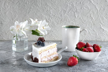 Piece of white mousse dessert cake with blueberries and chocolate on a plate with strawberries, cup of tea and white flowers on grey textured background. Beautiful breakfast. Selective focus