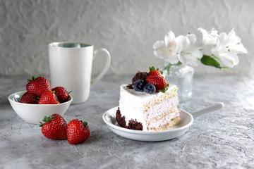 Piece of white mousse dessert cake with blueberries and chocolate on a plate with strawberries, cup of tea and white flowers on grey textured background. Beautiful breakfast. Selective focus
