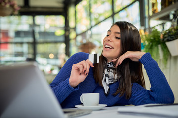 Attractive caucasian brunette in sweater sitting in cafe and holding credit card while looking at...