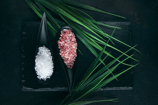 Pink Himalayas and white sea salt crystals in black spoons on stone plate with tropical plant. Dark background, top view. Food or spa concept.