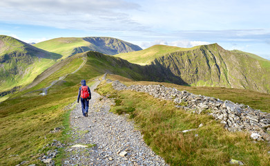 Fototapeta na wymiar A hiker walking along a stone chip path on a mountain ridge towards the summits of Hobcarton and Hopegill Head in the Lake District, England, UK.
