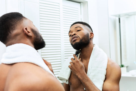 Young man shaving his beard with electric shaver, looking at mirror