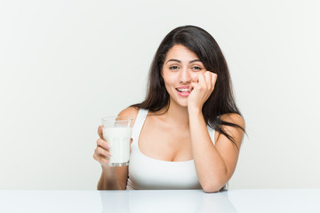 Young hispanic woman holding a glass of milk biting fingernails, nervous and very anxious.