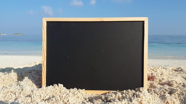 A blank black chalkboard rests on top of clean white sand on a tropical beach in the Caribbean